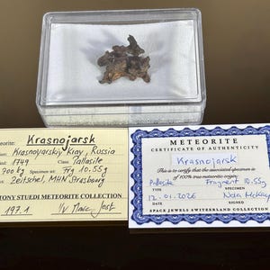 Puede incluir: Un fragmento de meteorito Krasnojarsk de 10,55 g, presentado en una caja transparente. Dos tarjetas proporcionan información sobre el origen y la autenticidad del meteorito. El meteorito es de tipo Pallasita. El texto es legible.