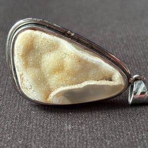 Puede incluir: Un colgante en forma de lágrima con una piedra druzy central de superficie cristalina y texturizada en color crema, engastada en un marco de metal plateado pulido con una anilla a juego fijada en el lateral para colgar.