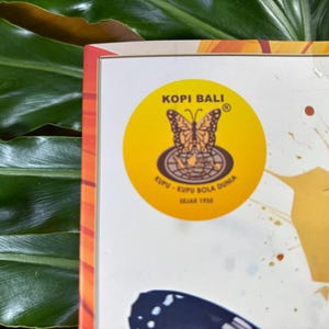 Puede incluir: Primer plano de una caja con una etiqueta circular amarilla con un dise&ntilde;o de mariposa y el texto "Kopi Bali". La etiqueta tambi&eacute;n incluye el texto "Kupu-Kupu Bola Dunia Sejak 1938". La caja est&aacute; sobre un fondo de hojas verdes.