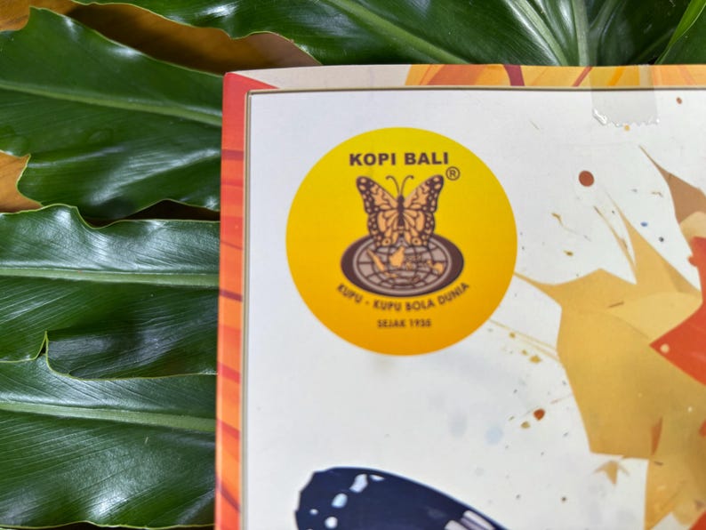 Puede incluir: Primer plano de una caja con una etiqueta circular amarilla con un dise&ntilde;o de mariposa y el texto "Kopi Bali". La etiqueta tambi&eacute;n incluye el texto "Kupu-Kupu Bola Dunia Sejak 1938". La caja est&aacute; sobre un fondo de hojas verdes.