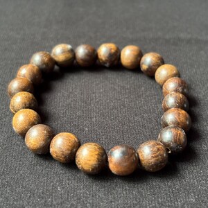 Puede incluir: Pulsera de cuentas de madera redondas, de color marrón oscuro. Cada cuenta tiene un patrón de vetas de madera natural. La pulsera se muestra sobre una superficie negra.