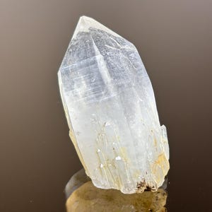 May include: A clear, crystalline quartz point with a faceted tip. The quartz is translucent with subtle internal striations and a light, natural color. The base of the crystal has a rough, earthy texture, and the background is a blurred brown.