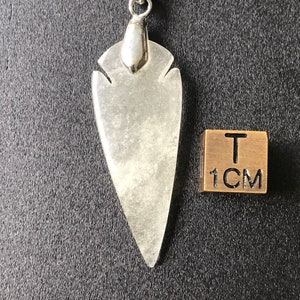 May include: A clear, teardrop-shaped pendant with a silver-colored bail hangs from a black cord. The pendant is next to a small, square, gold-colored block with the text "T" and "1CM" on it. The background is dark gray.