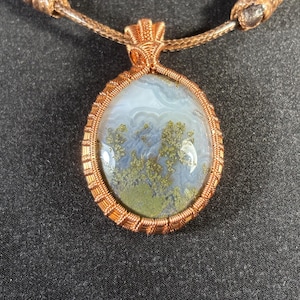 Könnte beinhalten: Ein Anhänger aus Kupferdraht, der einen Moosachat-Stein mit einem weißen und grünen Muster umwickelt. Der Anhänger ist an einer braunen Schnur befestigt.