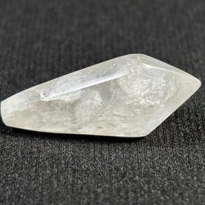 Può includere: Un cristallo trasparente e sfaccettato con un aspetto leggermente opaco. Il cristallo è allungato con un'estremità appuntita e poggia su una superficie scura e strutturata. La forma del cristallo è geometrica, riflettendo la luce.