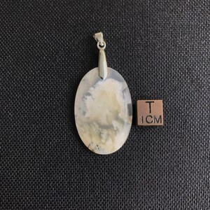 May include: An oval-shaped pendant with a silver bail. The pendant features a natural stone with a cloudy, cream and gray pattern. A 1 cm ruler is next to the pendant.