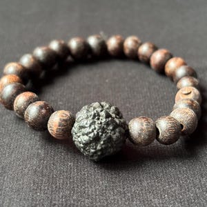 Puede incluir: Una pulsera de cuentas marrón oscuro con una cuenta central negra texturizada. La pulsera presenta cuentas redondas de madera con un patrón de vetas natural. Las cuentas están ensartadas, formando una forma circular.