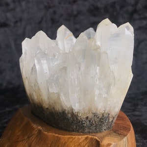 Pode incluir: Um aglomerado de cristais de quartzo transparentes sobre uma base de madeira. Os cristais estão dispostos em uma formação pontiaguda voltada para cima.