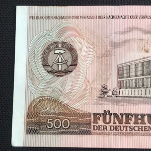 May include: A 500 East German Mark banknote featuring a building with a red roof and a compass and hammer symbol in a circle. The text "WER BANKNOTEN NACHMACHT ODER VERFALSCHT ODER NACHGEMACHTE ODER VERFALSCHTE SICH VERSCH" is printed at the top of the banknote. The text "F&Uuml;NFHUND" and "DER DEUTSCHEN DEMO" is printed at the bottom of the banknote.
