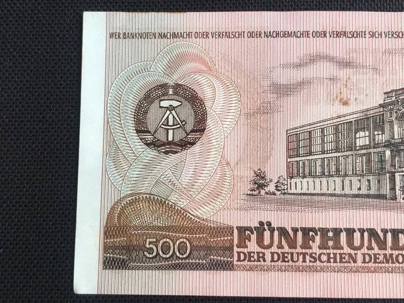May include: A 500 East German Mark banknote featuring a building with a red roof and a compass and hammer symbol in a circle. The text "WER BANKNOTEN NACHMACHT ODER VERFALSCHT ODER NACHGEMACHTE ODER VERFALSCHTE SICH VERSCH" is printed at the top of the banknote. The text "F&Uuml;NFHUND" and "DER DEUTSCHEN DEMO" is printed at the bottom of the banknote.