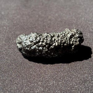 Pode incluir: Um objeto oblongo cinza escuro, texturizado, com uma superfície rugosa e porosa. O objeto projeta uma sombra sobre um fundo cinza escuro. A forma geral é irregular e orgânica.