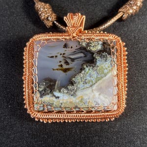 Puede incluir: Un colgante envuelto en alambre de cobre con una piedra de ágata cuadrada con patrones marrones, blancos y verdes. El colgante está unido a una cadena de cobre.