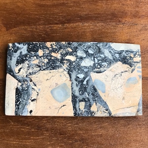 以下が含まれることがあります： 独特な大理石模様の研磨された長方形の石の板。石は主にベージュで、黒と灰色の筋と渦巻きがあります。