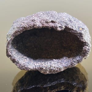 Op de afbeelding: Een donkergrijs, hol, rotsachtig object met een ruw, getextureerd oppervlak. Het object heeft een ovale vorm met een grote opening. De binnenkant is donker en contrasteert met de lichtere buitenkant. Het object wordt hieronder weerspiegeld.