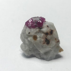 Peut inclure: Une petite roche blanche rugueuse avec un groupe de cristaux de rubis rouges sur le dessus. La roche contient des inclusions minérales brunes et jaunes.