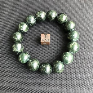 May include: A green and white speckled stone bracelet with 1 cm beads. The bracelet is laid out on a dark grey textured surface with a small silver cube that reads "1 CM" in the centre of the image.