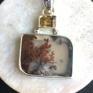 Puede incluir: Un colgante de plata con una piedra de ágata dendrítica rectangular, una pequeña piedra de topacio y una piedra de amatista púrpura.