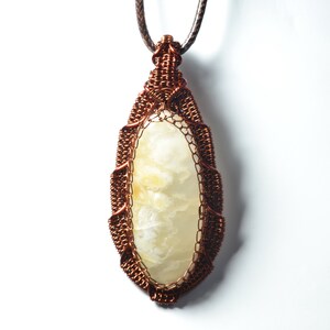Pode incluir: Um pingente envolto em fio de cobre com uma pedra de ágata branca. O pingente tem formato oval e pendura em um cordão marrom.