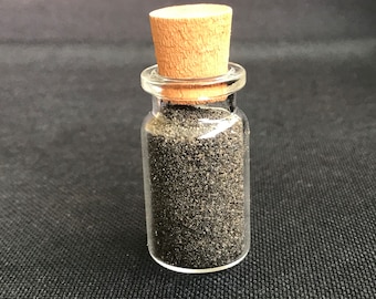 Puede incluir: Una pequeña botella de vidrio transparente llena de gránulos de color gris oscuro, sellada con un tapón de corcho. El contenido de la botella parece ser un material fino y granular. El fondo es una superficie oscura y texturizada.