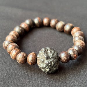 May include: A bracelet made of dark brown wooden beads and a textured, dark grey central bead. The bracelet is circular and sits on a dark background. The wooden beads have a natural grain.
