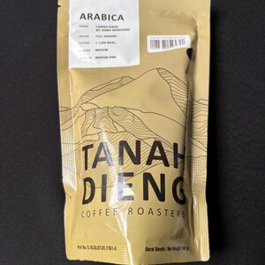 Op de afbeelding: Een goudkleurige koffiezak met de tekst "ARABICA" en "TANAH DIENG COFFEE ROASTERS". De zak bevat details over de herkomst, het proces, de hoogte en de maling van de koffie. Het nettogewicht is 100 g.