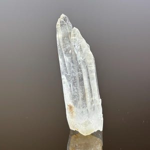 May include: A clear quartz crystal cluster with a natural, rough texture. The crystal is transparent with a slight milky hue and a pointed tip. The quartz is displayed against a dark background, highlighting its clarity.