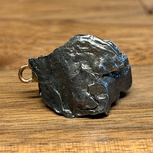 Puede incluir: Un pequeño meteorito de forma irregular, de color gris oscuro, con un lazo de metal dorado unido. El meteorito tiene una superficie rugosa y texturizada.