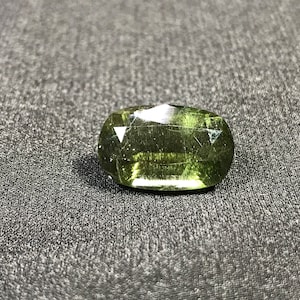May include: A green gemstone with a faceted cut. The stone has a slightly cloudy appearance and is set in a bezel setting.