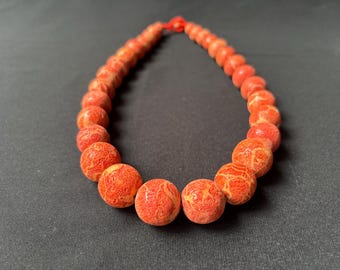 Könnte beinhalten: Eine Halskette mit runden, korallenfarbenen Perlen mit strukturierter Oberfläche. Die Perlen sind auf einer roten Schnur aufgefädelt und bilden ein auffälliges Accessoire. Die Halskette wird vor einem dunklen Hintergrund präsentiert. Die Perlen haben einen Durchmesser von ca. 1 cm.