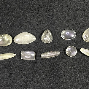 Puede incluir: Una colección de piedras preciosas claras y pulidas en varias formas, incluyendo ovaladas, de lágrima y rectangulares. Las piedras tienen un aspecto ligeramente moteado y están dispuestas sobre una superficie oscura.