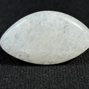 Può includere: Una gemma lucidata di forma ovale, dall'aspetto pallido e traslucido. La pietra ha una superficie liscia e arrotondata e una sottile trama interna. La gemma è posta su uno sfondo scuro e strutturato.