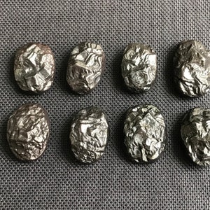 May include: Ten oval-shaped, dark gray, metallic stones with a rough, textured surface. The stones have a crystalline appearance.