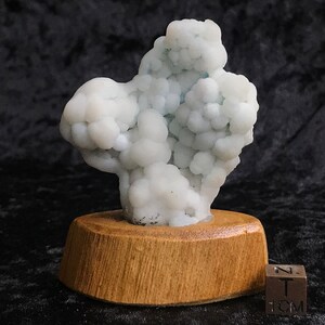 Pode incluir: Um espécime mineral branco e com bolhas em um suporte de exibição de madeira. O espécime tem aproximadamente 5 cm de altura. Uma pequena régua de metal está incluída para escala.