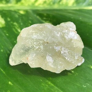 May include: A clear, translucent, irregularly shaped crystal with a slightly yellow hue. The crystal is resting on a green leaf.