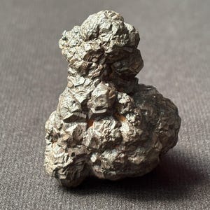 May include: A dark, textured mineral specimen with a rough, crystalline surface. The stone has a dark gray color and an irregular shape, with a cluster of protruding formations. The mineral sits on a gray surface.
