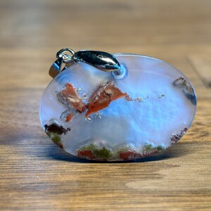 Puede incluir: Péndulo de forma ovalada hecho de ágata transparente con inclusiones marrones y verdes. El colgante tiene una anilla de plata.