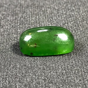 Könnte beinhalten: Ein glatter, oval geformter, grüner Edelstein. Die polierte Oberfläche reflektiert das Licht, kleine dunkle Einschlüsse sind sichtbar. Der Edelstein liegt auf einem strukturierten grauen Hintergrund, der seine lebendige Farbe und Form hervorhebt. Dies könnte ein Cabochon für Schmuck sein.