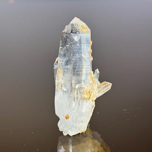 Może przedstawiać: Przezroczysty kryształ kwarcu ze spiczastym wierzchołkiem i wieloma mniejszymi formacjami kryształowymi. Kryształ ma półprzezroczysty wygląd z obszarami jasnobrązowych złóż mineralnych. Kryształ jest wyświetlany na ciemnym tle.