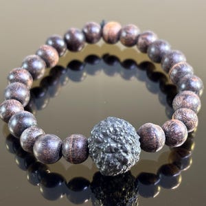May include: A bracelet with dark brown wooden beads and a textured, dark grey central bead. The beads are arranged on a band, displayed on a reflective surface, showing the wood grain and the unique design.