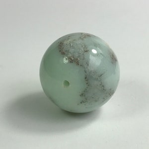 May include: A light green, round, smooth, polished stone bead with a small hole in the center. The bead has a mottled appearance with darker gray and brown markings.