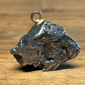 Puede incluir: Un colgante de meteorito de color plateado con un bucle dorado para colgar. El meteorito tiene una superficie rugosa e irregular.