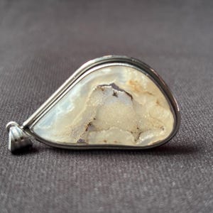 May include: A teardrop-shaped pendant featuring a polished silver-toned metal frame surrounding a translucent, off-white stone with a textured, crystalline druzy centre. The pendant includes a small metal bail at the narrow end for attachment.