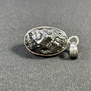 May include: A silver pendant featuring a dark gray, irregularly shaped meteorite encased in a silver oval setting. The pendant has a silver bail for attaching to a chain.
