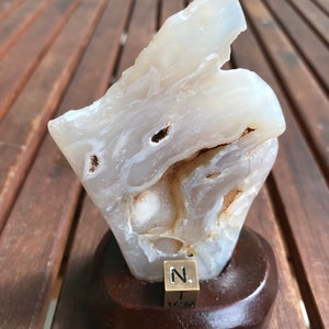 Puede incluir: Geoda de ágata blanca con bandas marrones y un centro hueco. La geoda se muestra en un soporte de madera con una placa de latón que dice "1 CM".