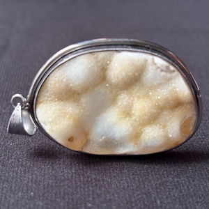 May include: An oval pendant featuring a textured, crystalline druzy stone in shades of cream, beige, and white. The stone is encased in a smooth, polished silver-toned metal bezel setting with a small circular bail attached to the left side.