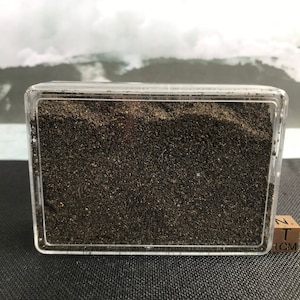 Pode incluir: Um recipiente de plástico transparente cheio de areia marrom escura. O recipiente está sobre uma superfície preta com um pequeno bloco de madeira que diz "1 CM" ao lado.