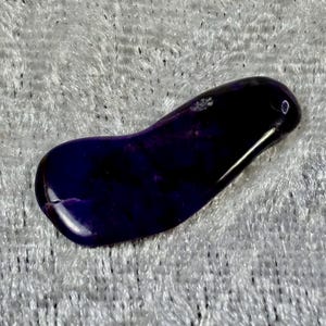 May include: A polished, teardrop-shaped stone with a deep purple hue. The stone has a smooth, glossy surface and appears to be made of a natural material. The color varies from dark to light purple, with some areas appearing almost black.
