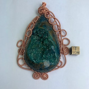 Puede incluir: Colgante artesanal con una piedra en forma de lágrima de color verde azulado y un interior brillante. Está encerrado en un intrincado trabajo de alambre de cobre con remolinos decorativos y una parte superior en forma de corona. Un cubo de 1 cm está al lado.