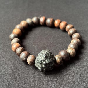 Könnte beinhalten: Ein Armband mit dunkelbraunen Holzperlen und einer zentralen, strukturierten, grauen Steinperle. Die Perlen sind auf einem Gummiband aufgefädelt. Die Holzperlen variieren von hell- bis dunkelbraun.