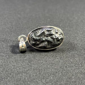 May include: A silver pendant featuring a black, oval-shaped meteorite with a rough, textured surface.